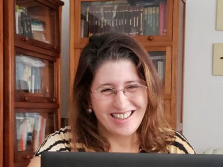 Mulher branca de cabelos castanhos pelo ombro, de óculos, sorrindo em frente a um computador